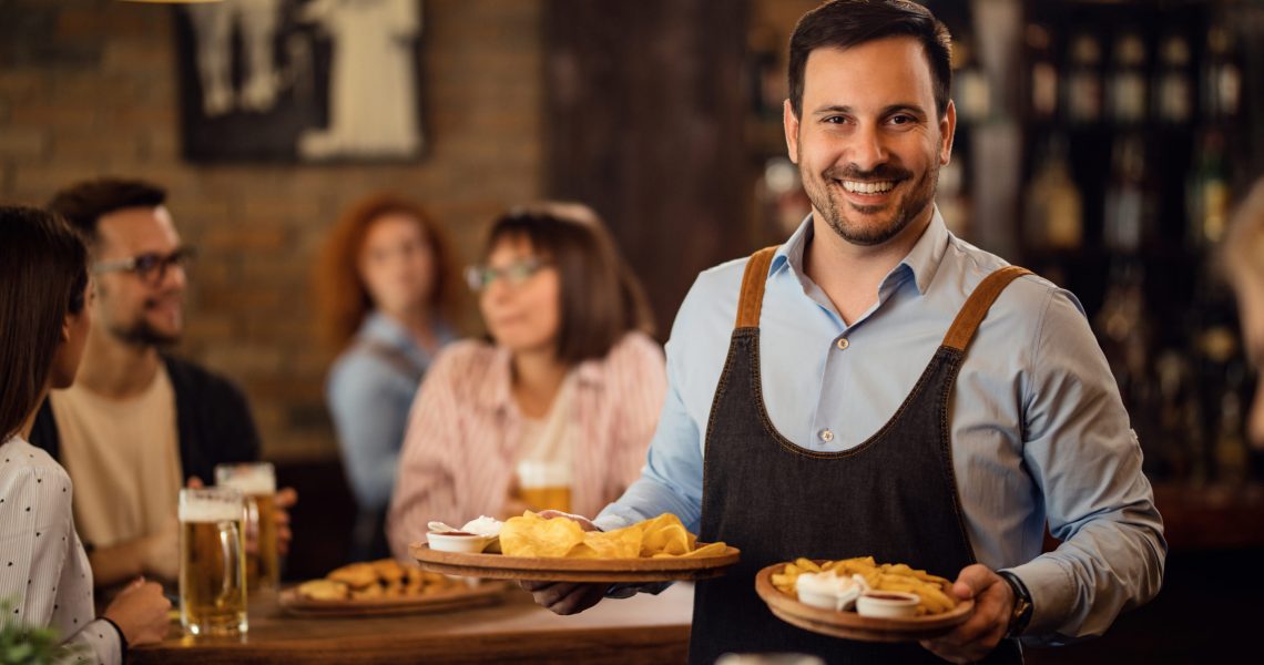 Happy waiter holding plates with food and looking at camera while serving guests in a restaurant.